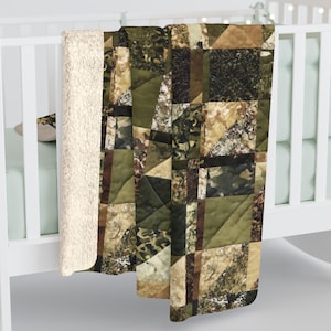 May include: A patchwork quilt with a camouflage pattern in shades of green, brown, and tan. The quilt is draped over a white crib, revealing a soft, cream-colored sherpa lining. The design features geometric shapes and textures.