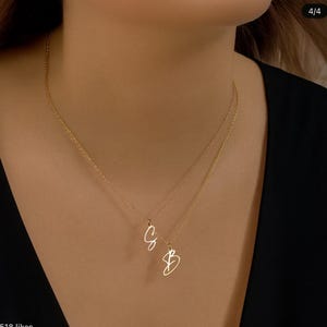 May include: A gold-colored necklace with two delicate chains. Each chain holds a cursive initial pendant, with the letters "C" and "B" visible. The necklace is worn against a dark background.
