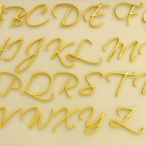 Könnte beinhalten: Ein vollständiges goldenes Alphabet von A bis Z in Schreibschrift. Die dreidimensionalen Buchstaben sind auf einem hellbeigen Hintergrund angeordnet und scheinen aus glänzendem Metall zu bestehen.