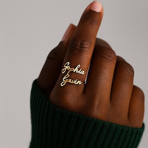 May include: A gold ring featuring the names "Sophia" and "Gavin" in a cursive font. The ring is displayed on a finger, set against a dark green sleeve. This is a personalised piece of jewellery.