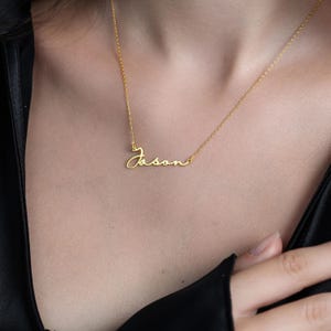 Puede incluir: Un delicado collar dorado con el nombre "Jason" escrito en cursiva. El collar se muestra sobre el pecho de una persona, con la cadena apoyada sobre la piel. El fondo es de color oscuro y sólido.