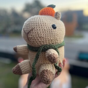 May include: A crocheted capybara plushie in shades of tan and brown, with a small orange pumpkin on its head and a green scarf around its neck. The plushie has black button eyes and is held in a hand.