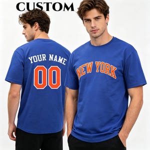 Könnte beinhalten: Ein königsblaues kurzärmeliges T-Shirt mit dem Text NEW YORK in Orange mit weißen Umrandungen auf der Vorderseite. Die Rückseite zeigt den Text YOUR NAME über der Nummer 00 in Orange mit weißen Umrandungen. Das Wort CUSTOM steht oben.