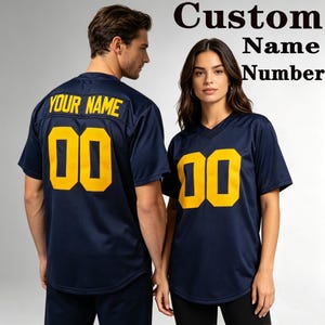 Pode incluir: Duas camisas esportivas azul-marinho de manga curta com decote em V. A parte de trás de uma camisa apresenta o texto amarelo YOUR NAME e o número 00, enquanto a frente da outra exibe o número amarelo 00. O texto diz Custom Name Number.