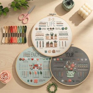 May include: Three embroidery hoops with various stitch examples and the words "LIFE" and "ENJOY!" are displayed. A collection of embroidery floss in various colors, pink scissors, and other decorative items are also present. The hoops are in wooden frames.