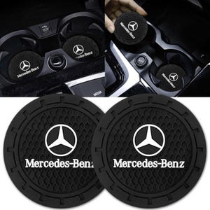 May include: Black car cup coasters with the Mercedes-Benz logo in white. The coasters are round and have a textured surface. The image shows the coasters in a car's cup holders and also a close-up of the coasters.