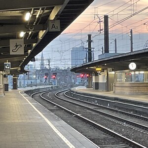 Könnte beinhalten: Ein Bahnhof in der Abenddämmerung, mit gekrümmten Gleisen, die sich vom Betrachter entfernen. Der Bahnsteig besteht aus Ziegeln, und es gibt metallische Überbauten. Der Himmel ist eine Mischung aus Rosa und Blau.