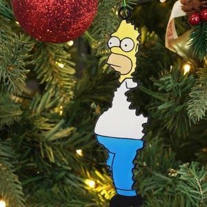 Puede incluir: Adorno navideño con Homer Simpson. El adorno es amarillo, con una camisa blanca y pantalones azules. Cuelga de un árbol de Navidad verde, junto a una bola roja brillante.