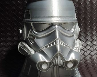 Puede incluir: Un enfriador de latas plateado con forma de casco de Stormtrooper de Star Wars. El enfriador tiene detalles del casco, incluyendo los ojos, la boca y las ventilaciones. Una lata plateada con texto verde y blanco está dentro.