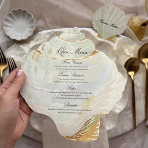 May include: A seashell-shaped menu card with the text "Our Menu" and a list of courses. The menu is white with gold accents and is held over a silver plate. Gold forks and spoons are also visible.