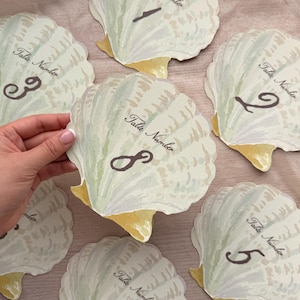 May include: A collection of seashell-shaped table number cards. Each card is pale green and white with a yellow base and a dark brown number. The words "Table Number" are printed on each card.