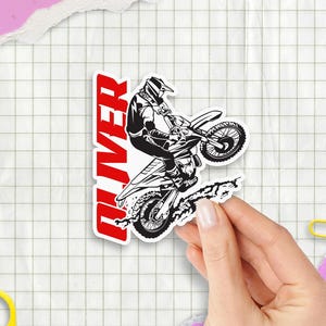 May include: A sticker featuring a black and white illustration of a motocross rider on a motorcycle, with the name "OLIVER" in red vertical text. The sticker is held against a grid paper background.