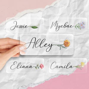 May include: Clear rectangular name tags with floral accents. Each tag features a name in elegant script: Jessie, Myebae, Alley, Eliana, and Camila. The tags are adorned with delicate illustrations of flowers, including lilies, daisies, marigolds, roses, and daffodils.