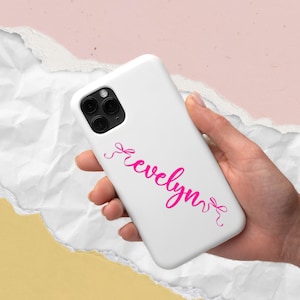 K&ouml;nnte beinhalten: Wei&szlig;es Handycase mit dem Namen "Evelyn" in rosa Schrift. Das Handycase hat eine Dreifach-Kamera. Der Hintergrund ist eine Mischung aus Wei&szlig;, Rosa und Gelb.