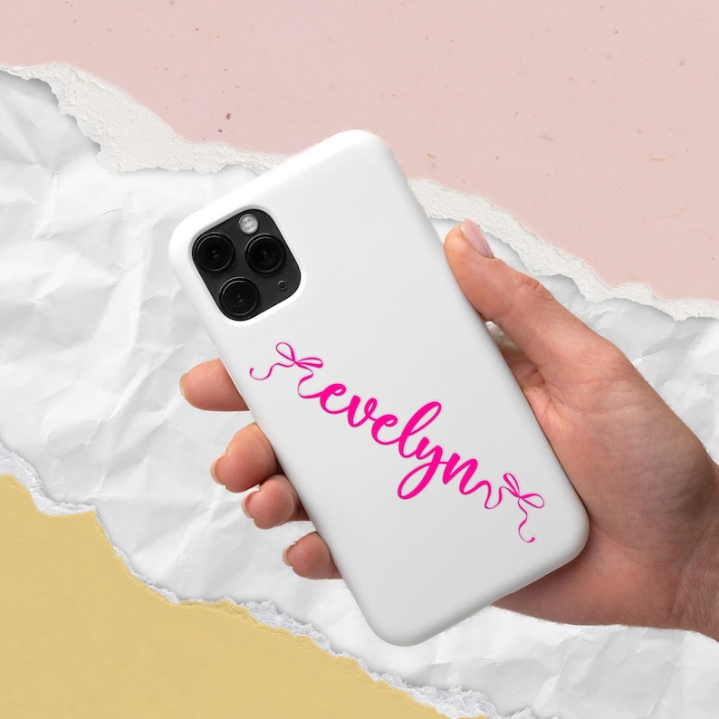 K&ouml;nnte beinhalten: Wei&szlig;es Handycase mit dem Namen "Evelyn" in rosa Schrift. Das Handycase hat eine Dreifach-Kamera. Der Hintergrund ist eine Mischung aus Wei&szlig;, Rosa und Gelb.