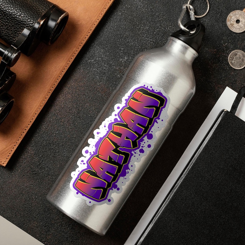 May include: A silver water bottle with a black cap and a keychain ring. The bottle is decorated with a sticker featuring the name "Nathan" in a graffiti-style design with red, purple, and yellow colors.