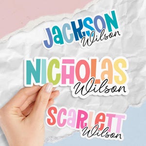 May include: Three colorful name stickers with the name "Jackson Wilson", "Nicholas Wilson", and "Scarlett Wilson". The names are in bold, bubble letters with a white outline. The background is white with torn paper and a pink and blue backdrop.