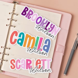 May include: Three colorful name stickers on a planner. The stickers read "Brooklyn Wilson", "Camila Wilson", and "Scarlett Wilson". The names are in various colors, including pink, orange, blue, and green. A silver pen rests on the planner.