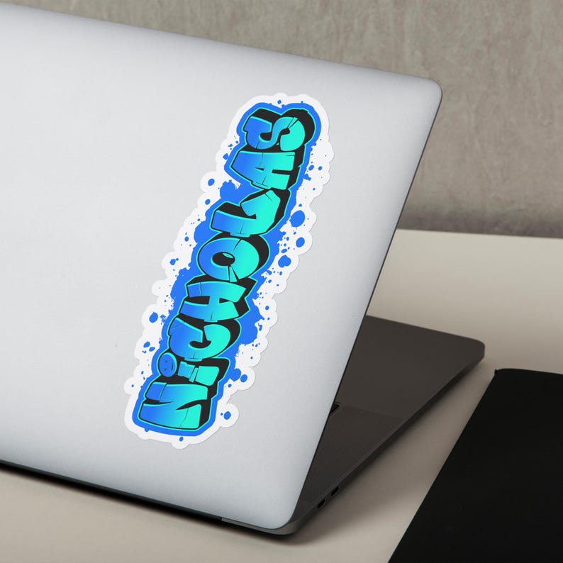 May include: A sticker with the name "Nicholas" in a graffiti-style font. The text is in shades of blue with a white outline and a blue splatter effect. The sticker is on a laptop.