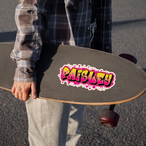 May include: A black skateboard with a colorful sticker that says "Paisley" in a graffiti style. The sticker has a yellow and pink gradient with white accents and pink splatters. The skateboard is held by a person wearing a plaid shirt.