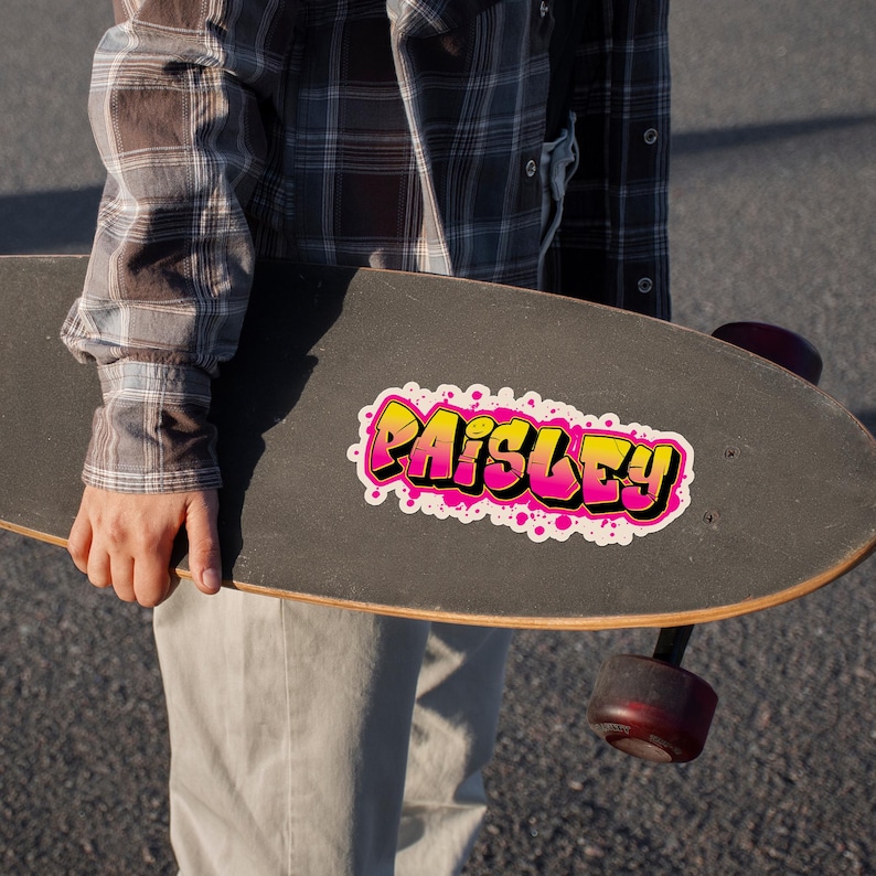 May include: A black skateboard with a colorful sticker that says "Paisley" in a graffiti style. The sticker has a yellow and pink gradient with white accents and pink splatters. The skateboard is held by a person wearing a plaid shirt.