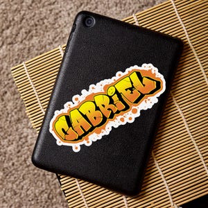 May include: A black tablet with a sticker that says "GABRIEL" in yellow and orange graffiti style lettering. The sticker has a white border and is placed on the tablet's surface.
