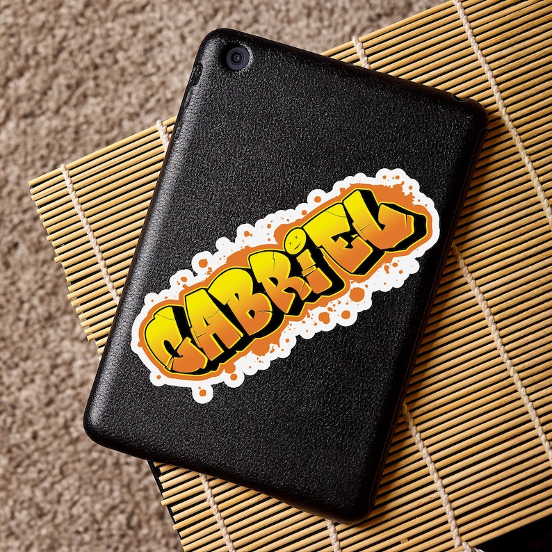 May include: A black tablet with a sticker that says "GABRIEL" in yellow and orange graffiti style lettering. The sticker has a white border and is placed on the tablet's surface.