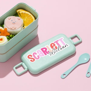 May include: A light green bento box with a matching lid featuring the name "Scarlett Wilson" in pink and red lettering. The box contains a pig-shaped rice ball, orange slice, and other food items. A light blue spoon is also visible.