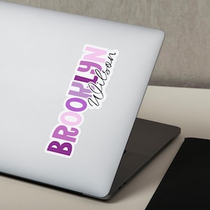 May include: A sticker with the name "Brooklyn Wilson" in a vertical layout. The word "Brooklyn" is in a gradient of purple hues, while "Wilson" is in a cursive font. The sticker is placed on a silver laptop.