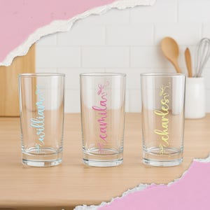 K&ouml;nnte beinhalten: Drei transparente Trinkgl&auml;ser mit Namen in farbiger Schrift: "William" in Blau, "Camila" in Pink und "Charles" in Gelb. Die Gl&auml;ser stehen auf einer Holzoberfl&auml;che, im Hintergrund K&uuml;chenutensilien.