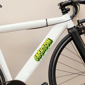 May include: Close-up of a white bicycle frame with a black wheel and tire. A green and yellow sticker with the name "JACKSON" is affixed to the frame. The bicycle has black handlebars and a black seat post.
