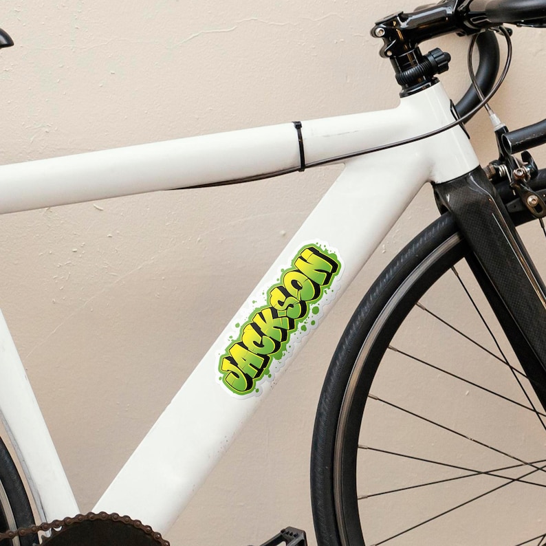 May include: Close-up of a white bicycle frame with a black wheel and tire. A green and yellow sticker with the name "JACKSON" is affixed to the frame. The bicycle has black handlebars and a black seat post.