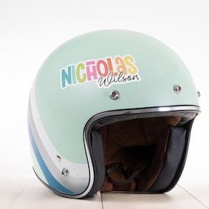 May include: A mint green motorcycle helmet with a retro design. The helmet features a colorful sticker with the name "Nicholas Wilson". A stripe design in blue, grey, and white accents the side. The interior is lined with brown fabric and black padding.