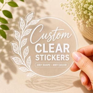 May include: A round, transparent sticker featuring white text that reads Custom CLEAR STICKERS and ANY SHAPE - ANY COLOR. The design includes a white line-art illustration of a leafy branch curving along the left side of the circular border.