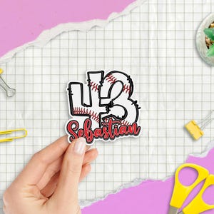 May include: A white sticker with the number 43 in a baseball design, with the name "Sebastian" in red script. The sticker is held by a hand against a grid paper background.