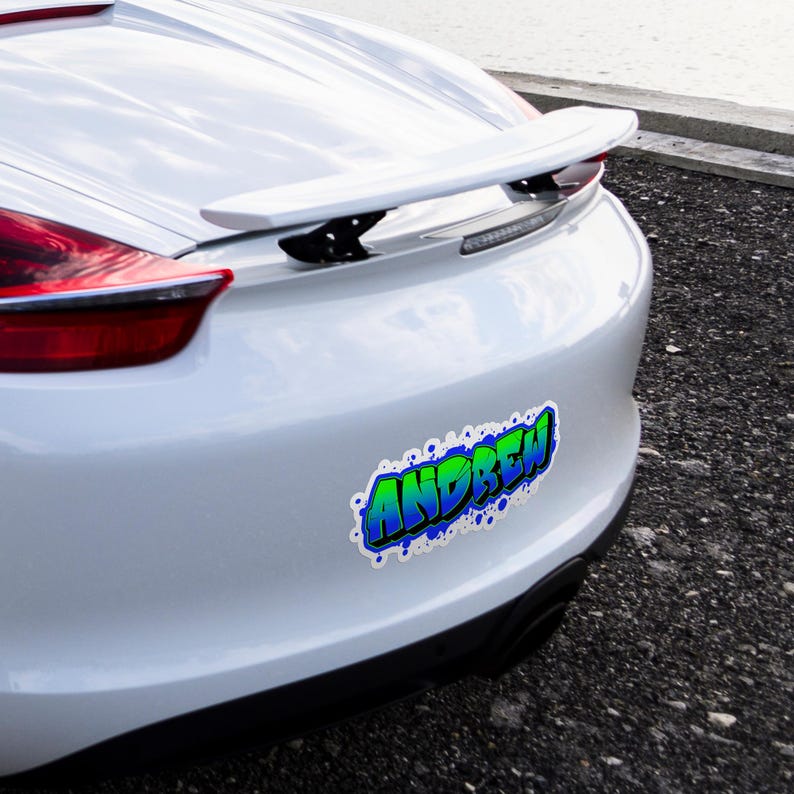 May include: A white car's rear bumper with a sticker that reads "ANDREW" in a graffiti-style font. The text is green and blue with a white outline and blue splatter accents.