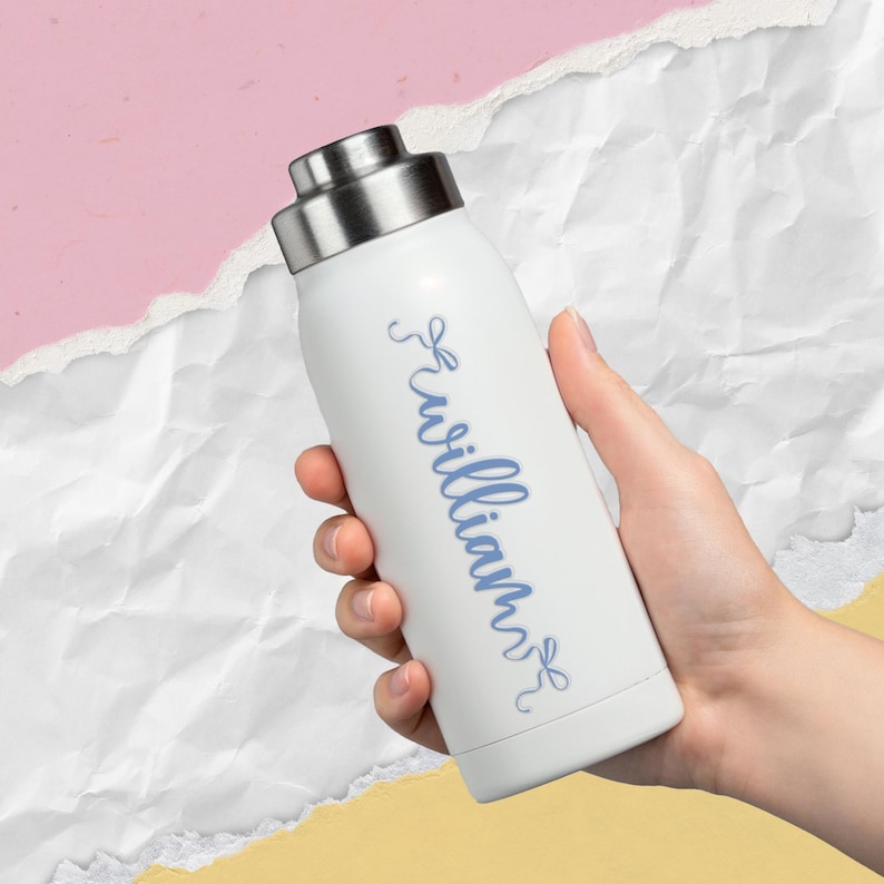 K&ouml;nnte beinhalten: Wei&szlig;e, isolierte Wasserflasche mit silbernem Deckel. Auf der Flasche steht der Name "William" in blauer Schrift. Die Flasche wird in einer Hand vor einem rosa, wei&szlig;en und gelben Hintergrund gehalten.