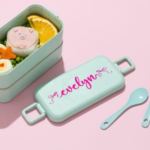 K&ouml;nnte beinhalten: Eine hellgr&uuml;ne Bento-Box mit passendem Deckel und L&ouml;ffel, vor rosa Hintergrund. Der Deckel tr&auml;gt den Namen "evelyn" in rosa Schrift. Die Bento-Box enth&auml;lt einen reisball in Schweineform, eine Orangenscheibe und andere Lebensmittel.