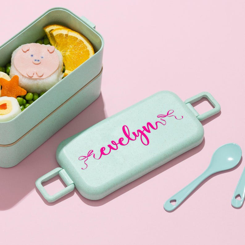 K&ouml;nnte beinhalten: Eine hellgr&uuml;ne Bento-Box mit passendem Deckel und L&ouml;ffel, vor rosa Hintergrund. Der Deckel tr&auml;gt den Namen "evelyn" in rosa Schrift. Die Bento-Box enth&auml;lt einen reisball in Schweineform, eine Orangenscheibe und andere Lebensmittel.