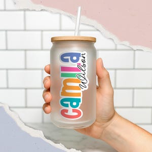 May include: Frosted glass tumbler with a wooden lid and a white straw. The tumbler features the name "Camila Wilson" in colorful, stacked letters. The glass is held by a person.