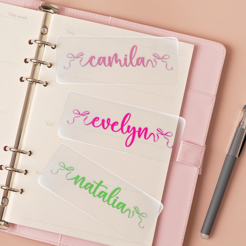 K&ouml;nnte beinhalten: Drei transparente Planer-Trenner mit Namen in Schreibschrift: "camila" in Pink und Lila, "evelyn" in leuchtendem Pink und "natalia" in Gr&uuml;n. Jeder Name hat eine Schleife. Die Trenner befinden sich in einem rosa Planer.