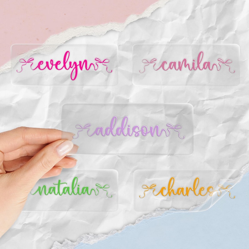 K&ouml;nnte beinhalten: Klare Namensschilder aus Acryl mit Namen in verschiedenen Farben: Evelyn, Camila, Addison, Natalia und Charles. Jedes Schild hat ein dekoratives Schleifendesign. Die Schilder werden auf einer wei&szlig;en, strukturierten Oberfl&auml;che angezeigt.