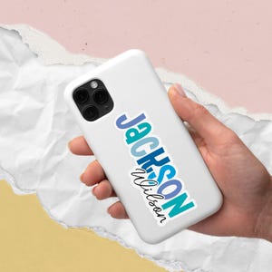 May include: A white smartphone case with a sticker that reads "Jackson Wilson" in blue and teal lettering. The phone has a triple-lens camera and is held in a person's hand against a colorful background.