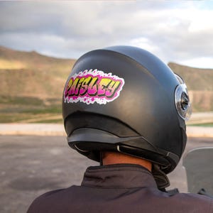 May include: Black motorcycle helmet with a sticker that says "Paisley" in pink and yellow graffiti-style lettering. The helmet has a clear visor and a dark neck guard. The background shows a mountain range and a cloudy sky.