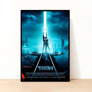 以下が含まれることがあります： 映画「トロン：レガシー」のフレーム入りポスター。青色の未来都市が描かれています。2人の人物がプラットフォームに立ち、光のビームに向かって腕を上げています。「TRON LEGACY」のタイトルが大きく表示されています。