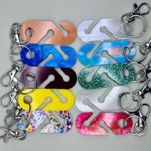 May include: A collection of colorful acrylic keychains with silver-tone hardware. The keychains feature a unique S-shaped design and are available in various colors and patterns, including glitter, marble, and solid hues. Each keychain has a clasp and a split ring.