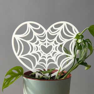 May include: A white heart-shaped plant trellis featuring a spider web pattern with a small heart cutout in the center. The trellis is inserted into a light green pot containing a green leafy plant with several perforated leaves.