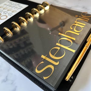 May include: A black planner with gold detailing, including the name "Stephanie" in gold script. The planner has gold disc binding and a gold pen. A sheet of stickers is visible in the upper left corner.