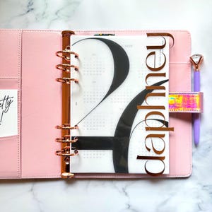 May include: A pink planner with copper-coloured hardware, open to a 2024 calendar page. The calendar page has a large black number 2 design. A purple pen with a diamond-shaped top rests in the planner's pen loop. The word "daphneu" is printed on the planner.