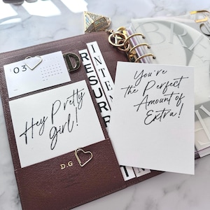May include: A brown leather planner with gold accents, holding cards with the phrases "Hey Pretty girl!" and "You're the Perfect Amount of Extra!" A small calendar and heart-shaped paper clips are also visible.